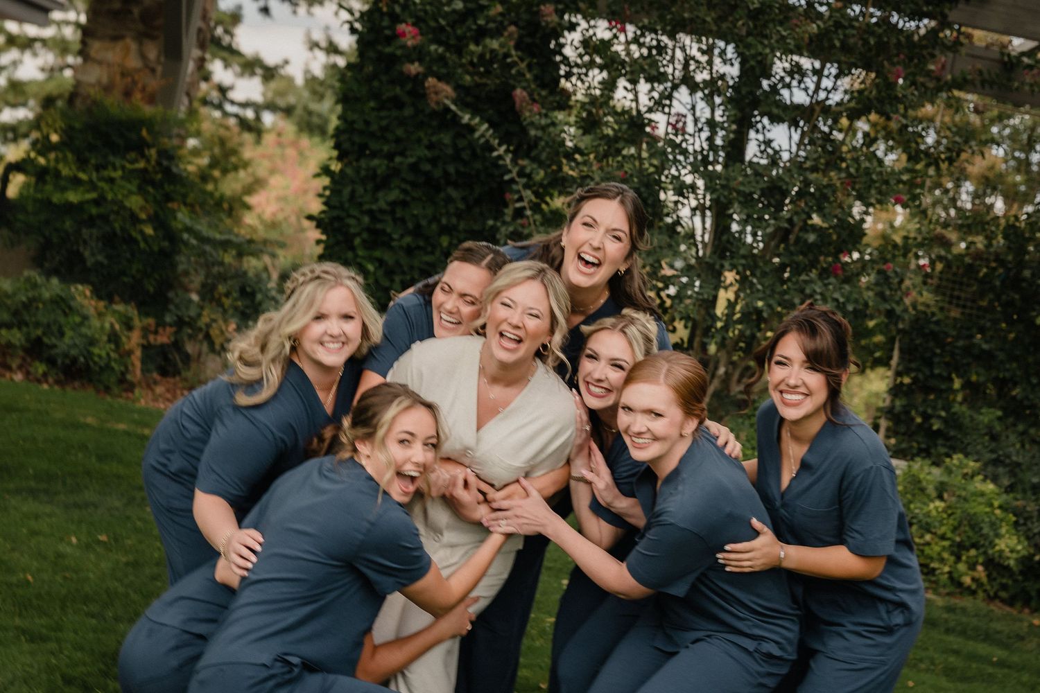 Wedding party laughing together outdoors in a garden setting during golden hour.