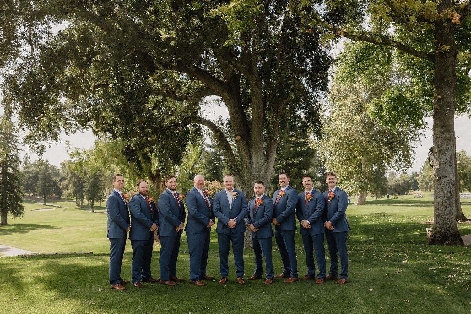 Wedding party in navy suits and dresses poses together on a sunny lawn with mature trees.