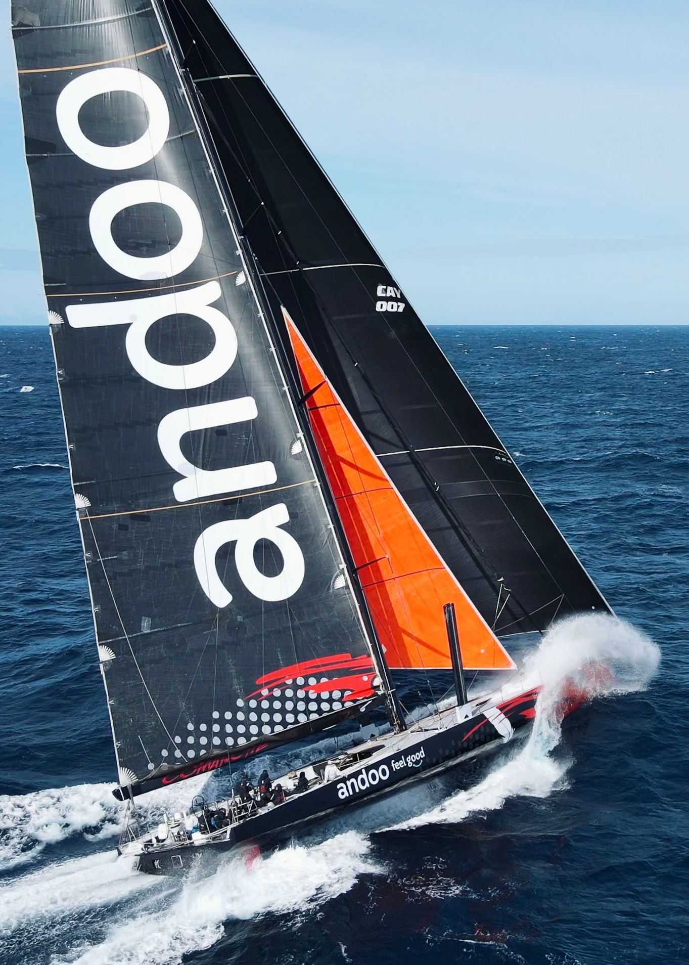 Racing yacht with black and orange sails cutting through ocean waves at high speed.