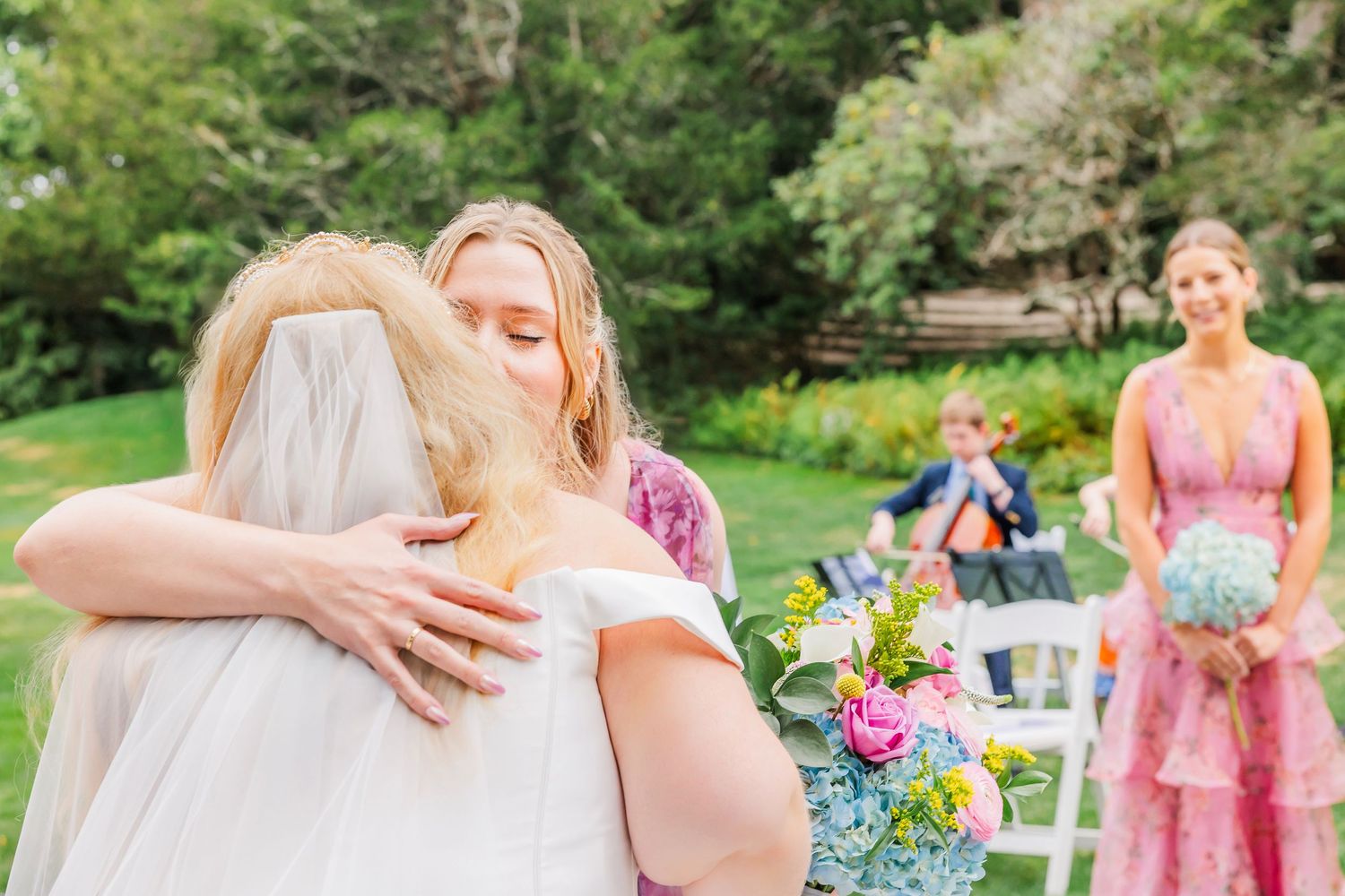 Bridesmaids in pink dresses embrace at an outdoor garden wedding celebration with colorful floral arrangements.