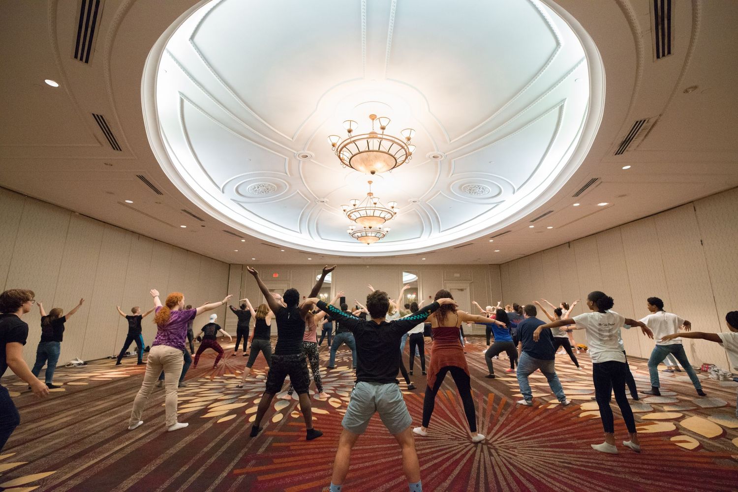 Large group fitness class doing stretching exercises in elegant ballroom with ornate chandelier and patterned carpet. Best photographer