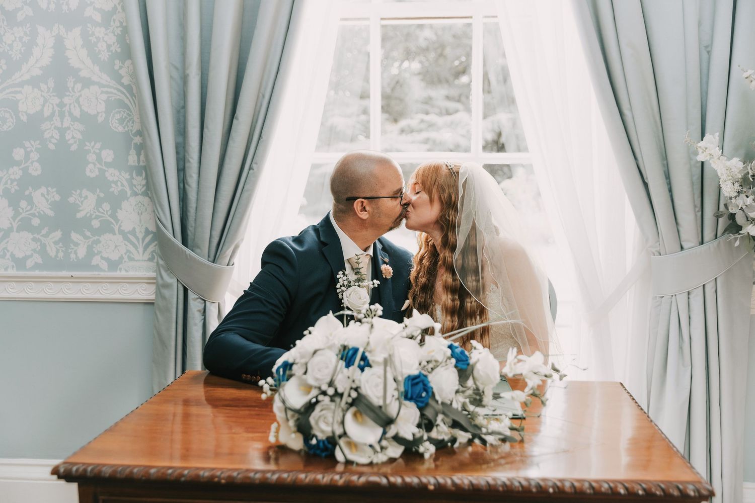 Bride and groom share a kiss at a wooden table with blue and white floral bouquet in the elegant blue room at Langtons House Registry