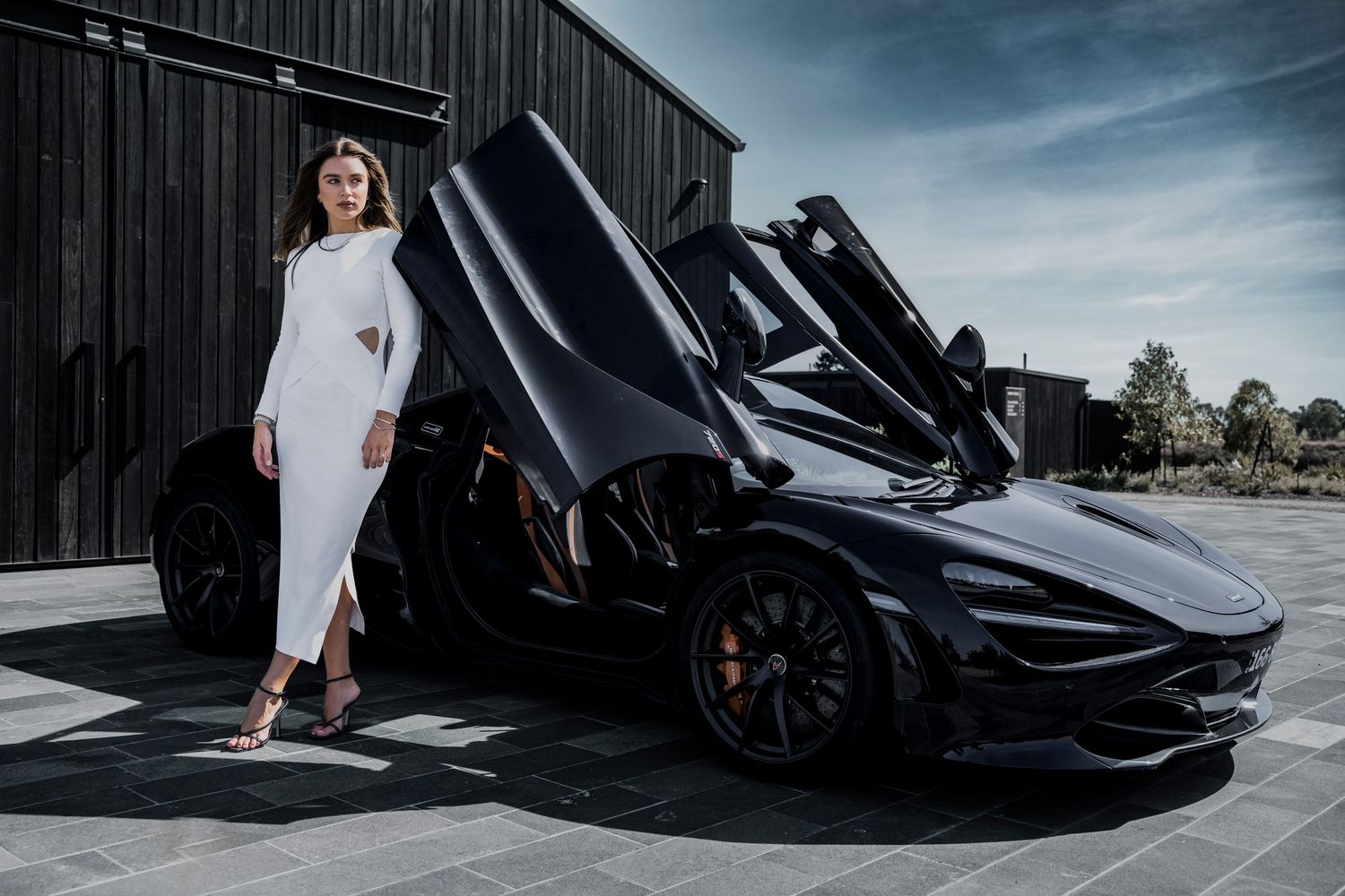Sleek black McLaren 720S supercar with dramatic butterfly doors open parked next to modern building at dusk.