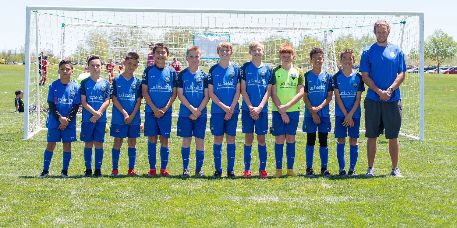 Denver Youth soccer team in blue uniforms lines up for team photo on sunny grass field with goal in background.