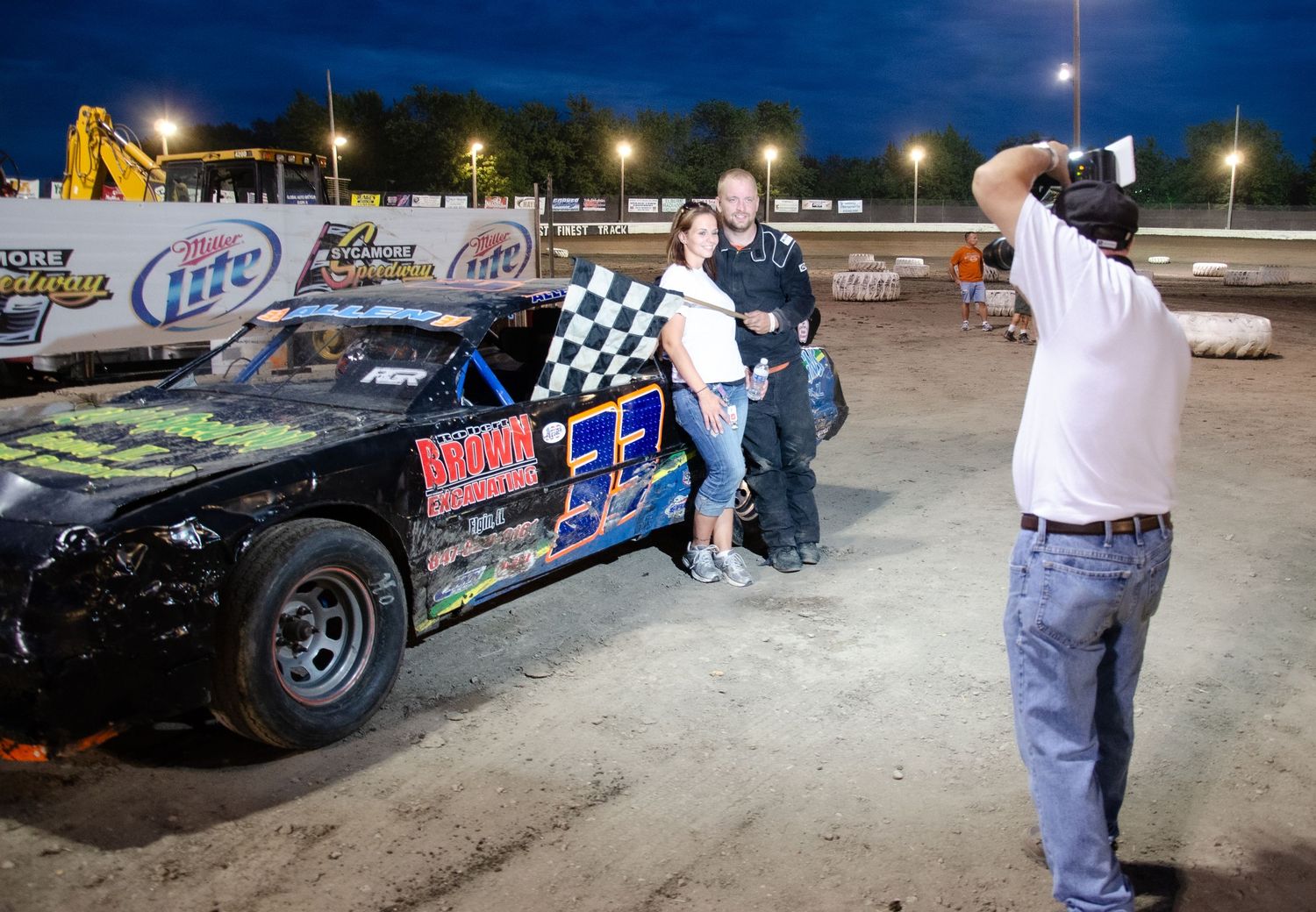 Race cars lined up at a nighttime racing event with crews working on vehicles under bright track lighting.