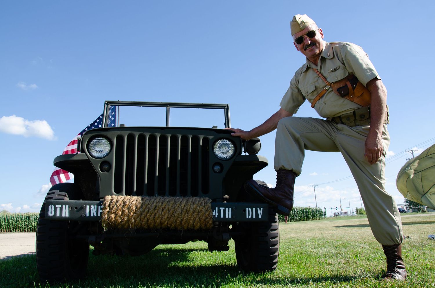 A vintage military Jeep is shown from a low angle on a grassy field against a blue sky background with a reenactor leaned against it.