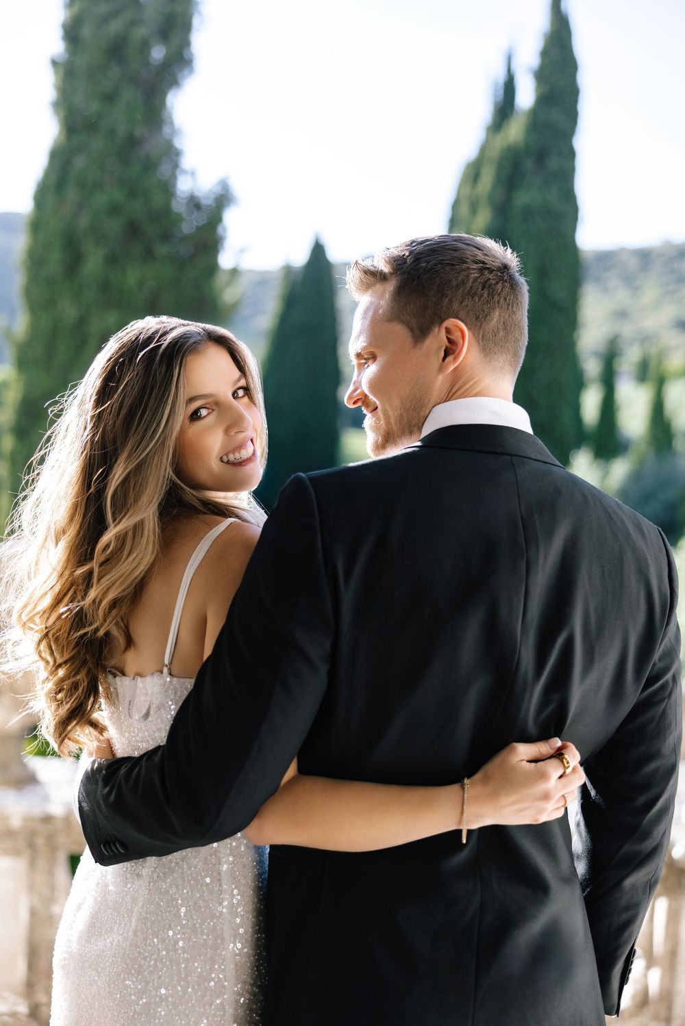 A couple in formal attire embracing outdoors with cypress trees in the background.