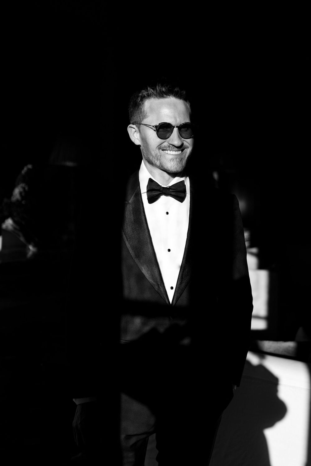 Black and white portrait of a man in tuxedo and sunglasses smiling.