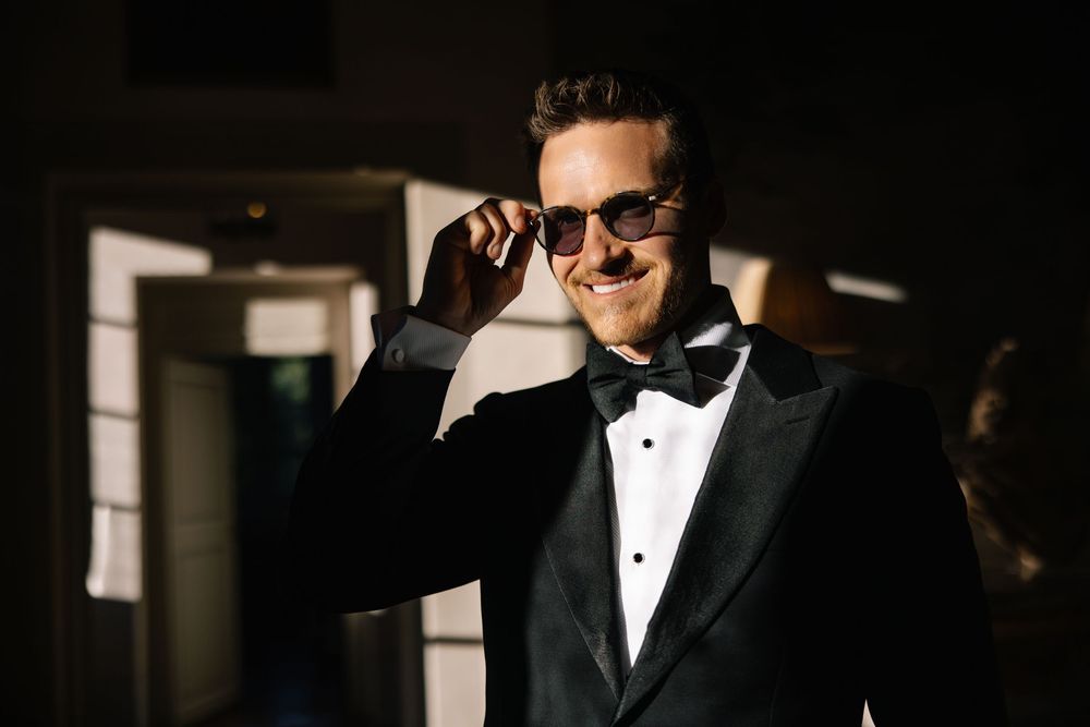 Groom in tuxedo and sunglasses poses dramatically in dark lighting.