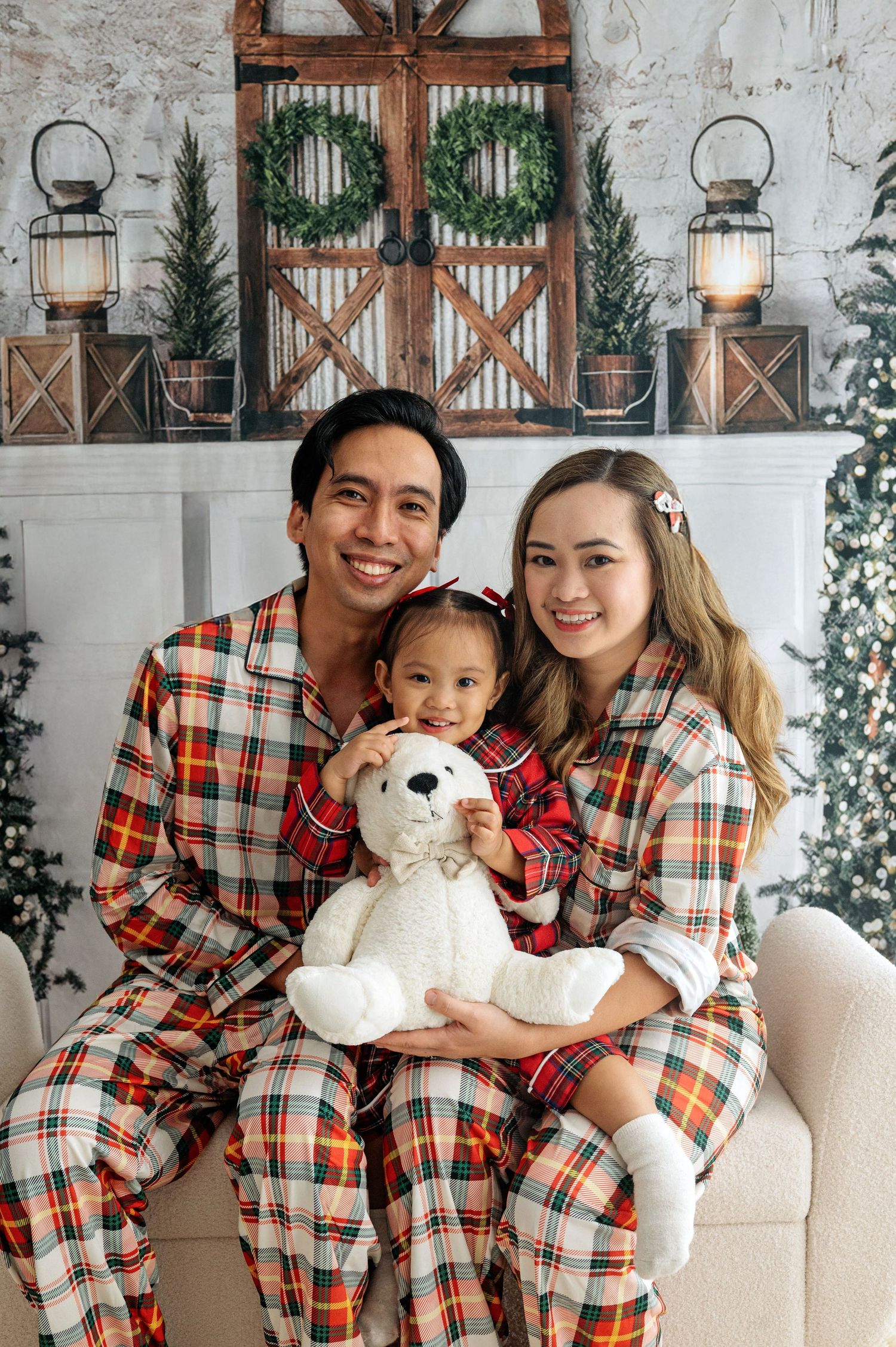 Family wearing coordinated plaid pajamas poses together with white teddy bear on cozy white rug for holiday photos.