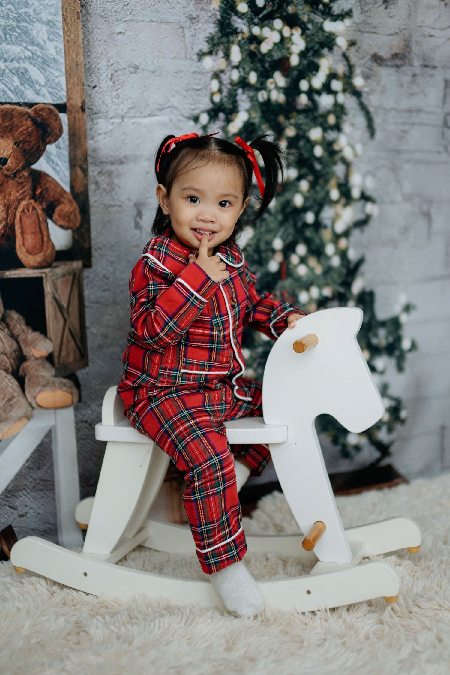 A child in a red plaid holiday outfit sits on a white rocking horse with Christmas decor in the background.