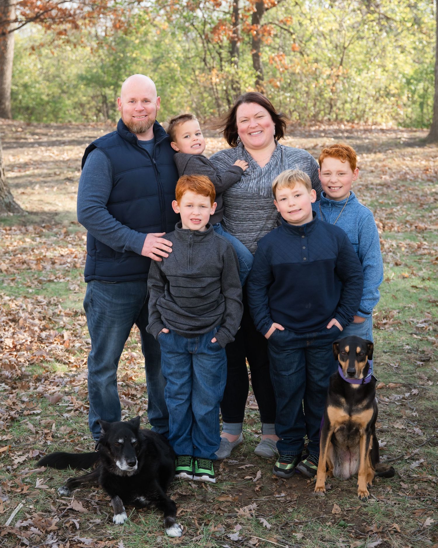 A family poses together on fallen autumn leaves with their loyal Rottweiler dog in an outdoor setting.