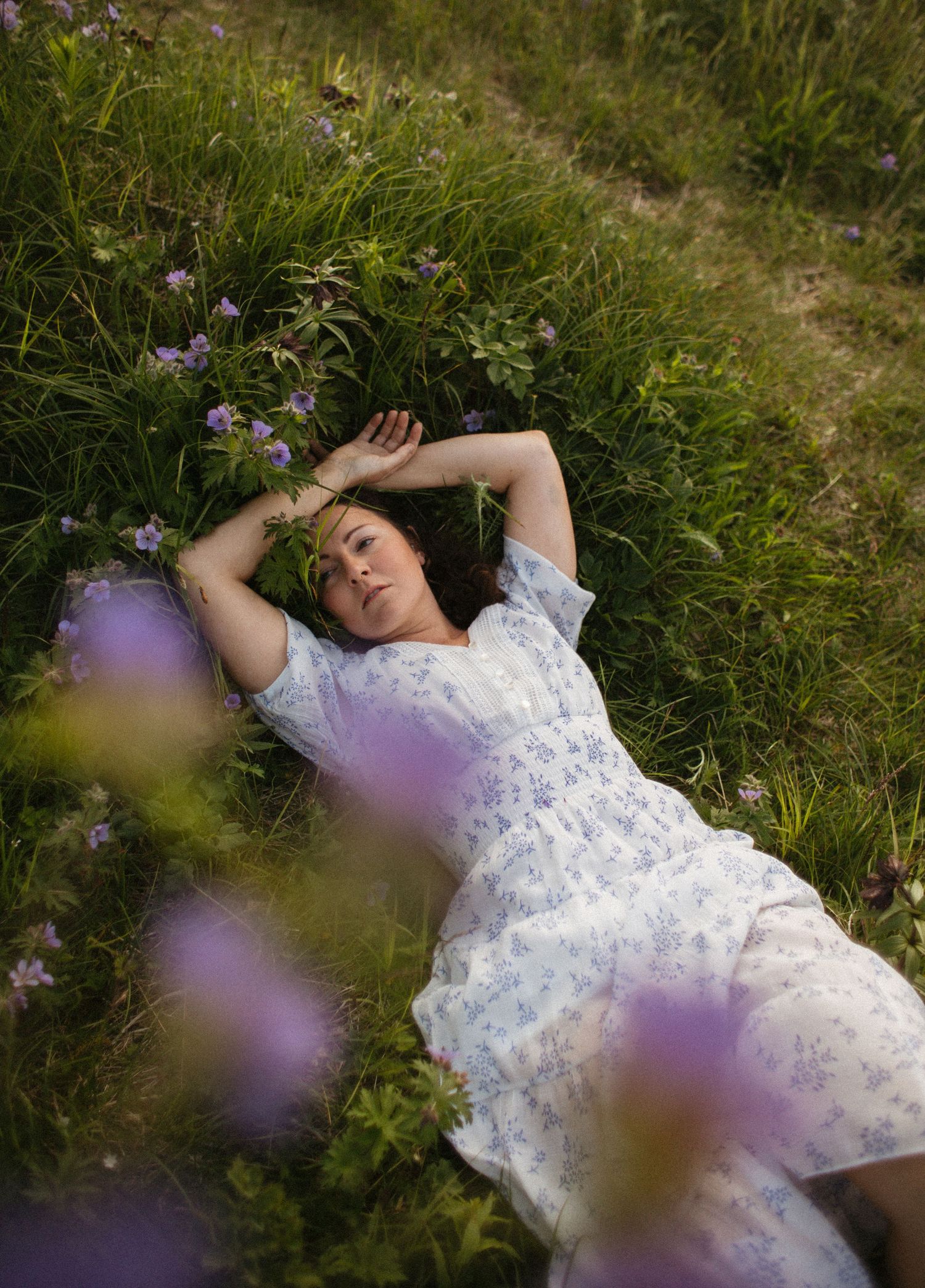 A person in a white floral dress lies peacefully in a meadow surrounded by purple wildflowers on a summer day.