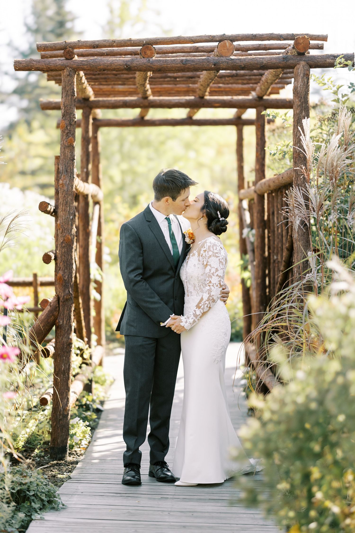 A couple shares an intimate moment under a rustic wooden pergola during their outdoor wedding ceremony.
