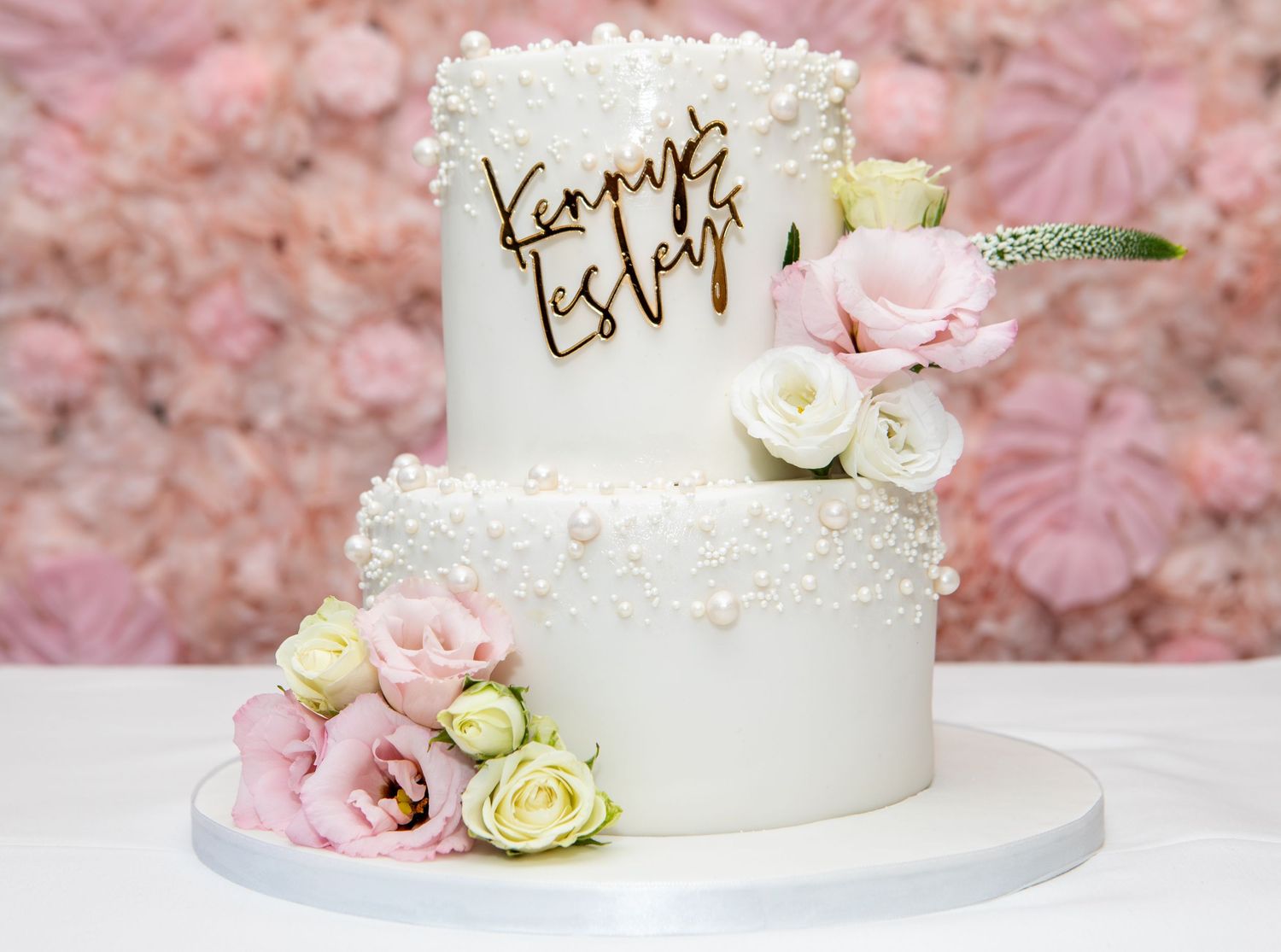 White wedding cake decorated with pink and cream roses on a pink floral background.
