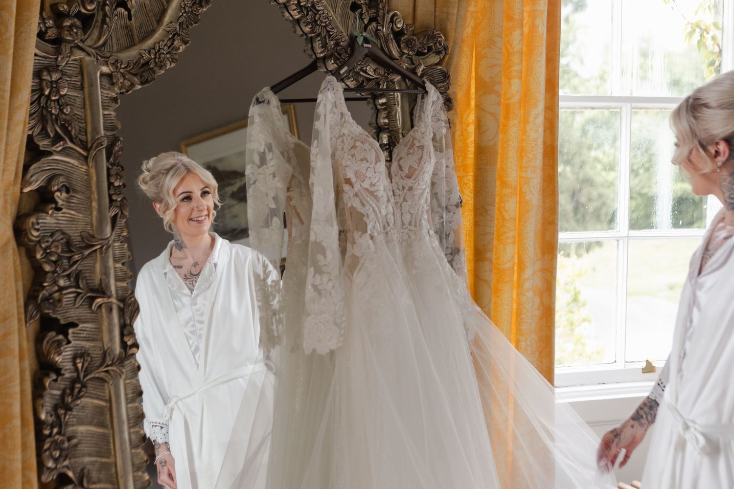 Bride in white robe admires wedding dress hanging by window, reflected in ornate gold mirror.