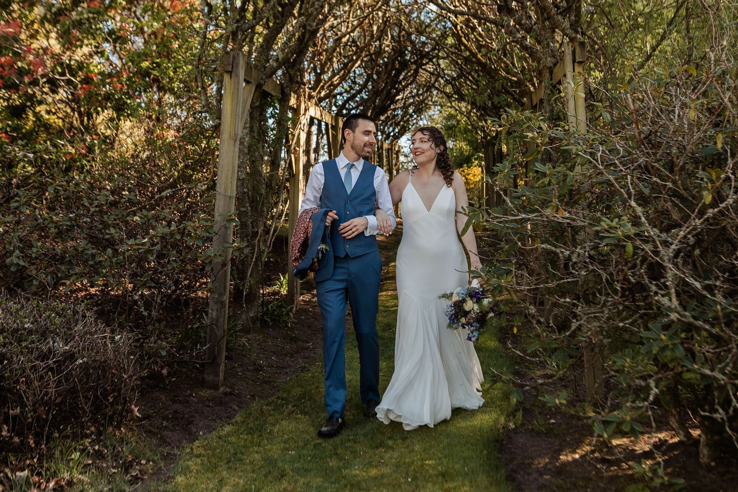 Bride and groom walking hand in hand through a lush garden archway on their wedding day.