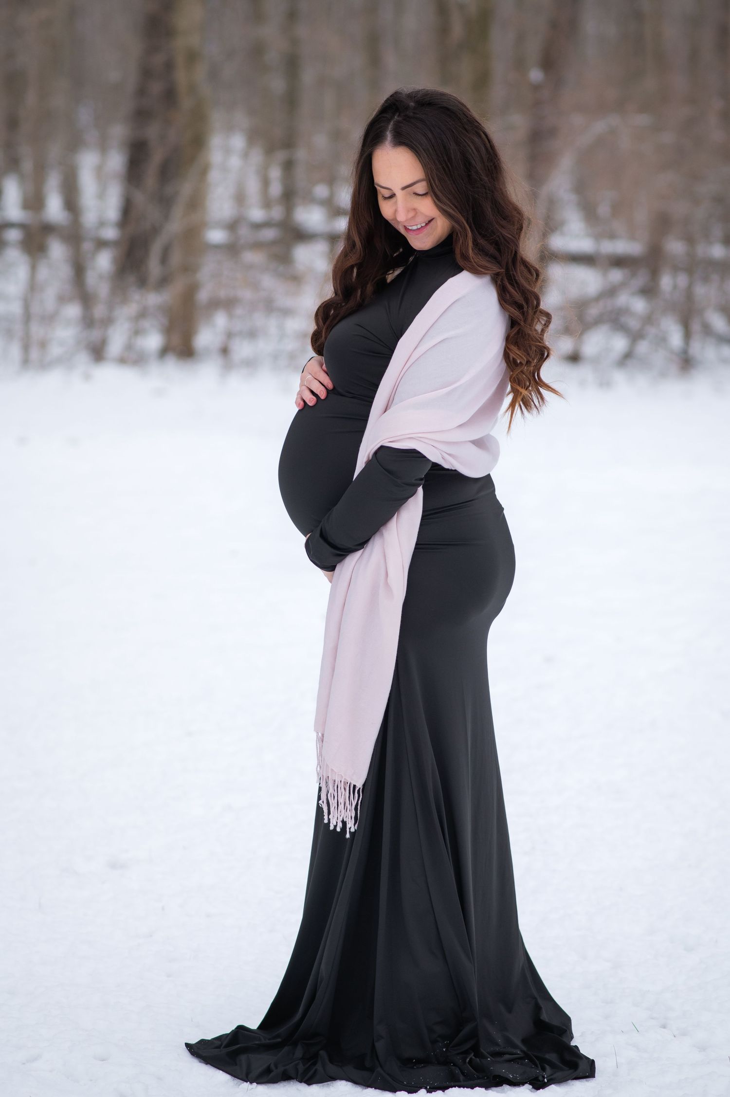 A person in a flowing black dress and light pink scarf poses gracefully in the snow during a winter photoshoot.