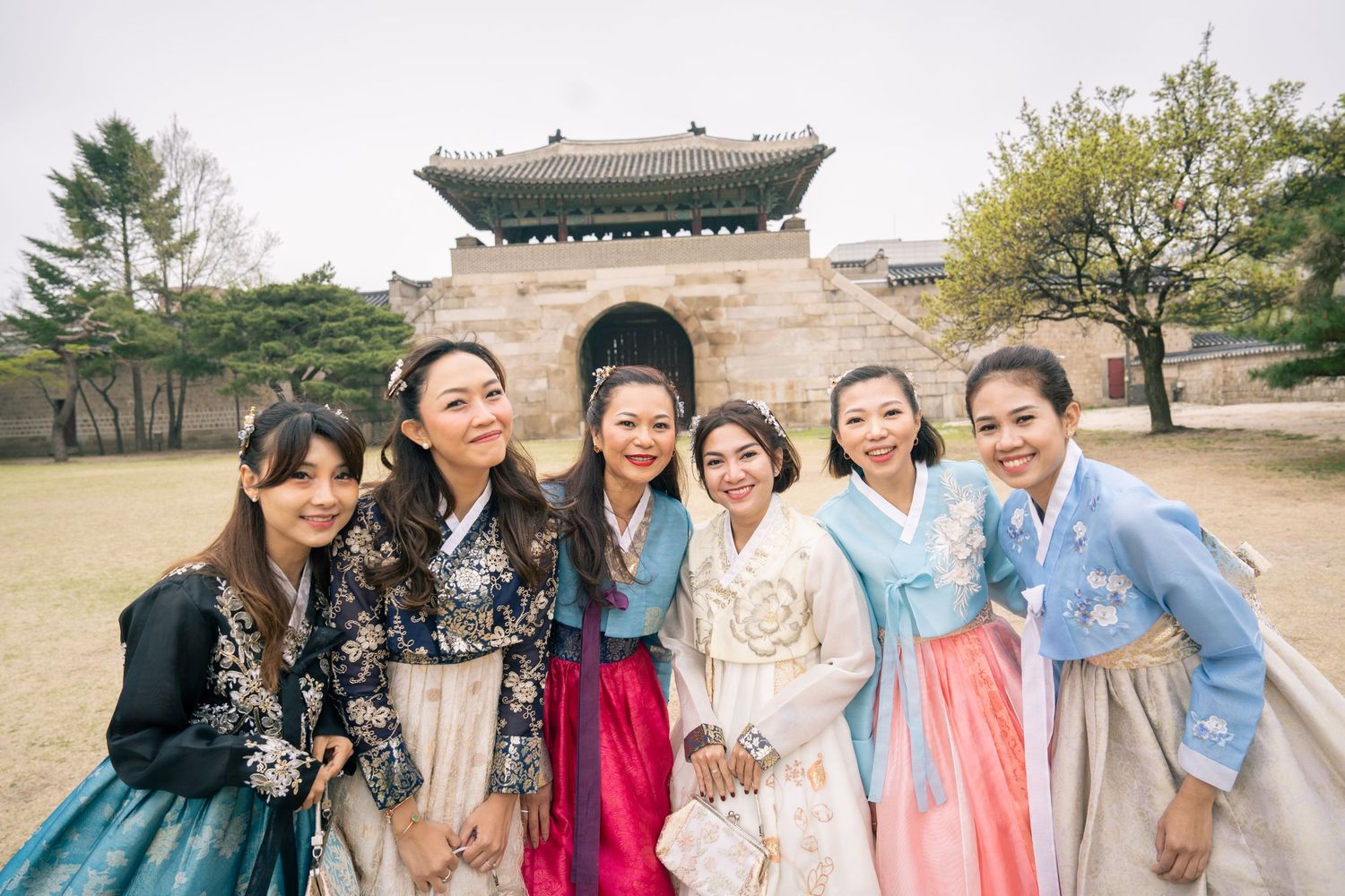A group wearing colorful traditional Korean hanbok dresses pose together in front of historic architecture.