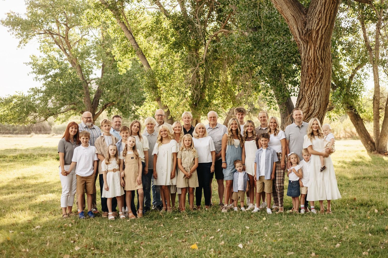 Large family group wearing coordinated white and beige outfits poses together in a sunlit park setting during golden hour.