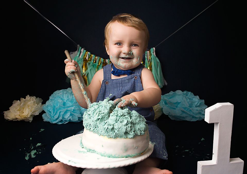 A baby in blue overalls sits in front of a turquoise cake with decorative tassels in the background.