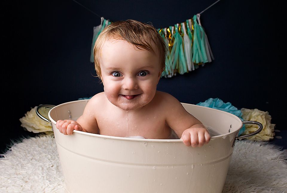 A smiling baby sits in a white basin with teal decorative bunting hanging in the background.