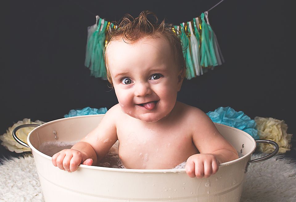 A baby sits in a white bathtub with blue and teal decorative tassels hanging in the background.