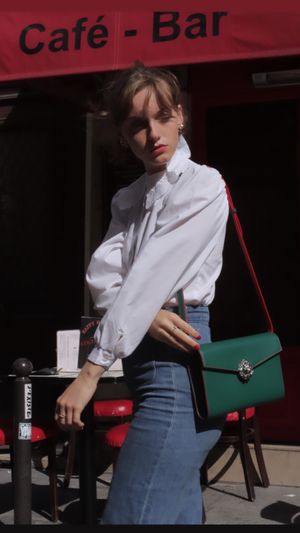 Fashion model wearing white button-up shirt and denim skirt with green handbag at outdoor cafe setting.
