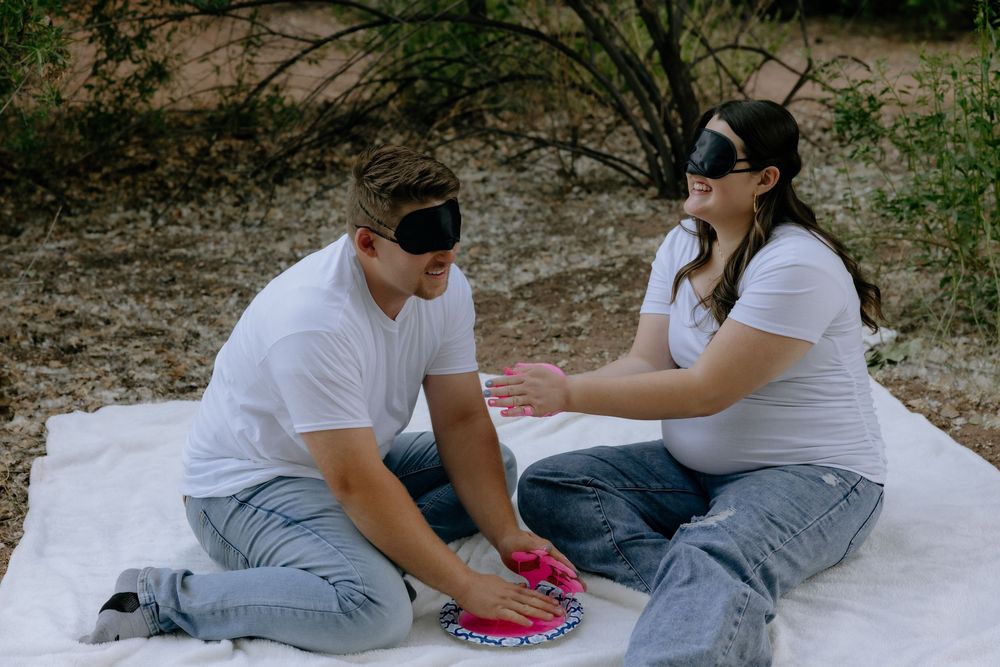 Two people in white shirts and jeans sit on a blanket outdoors playing cards while wearing blindfolds.