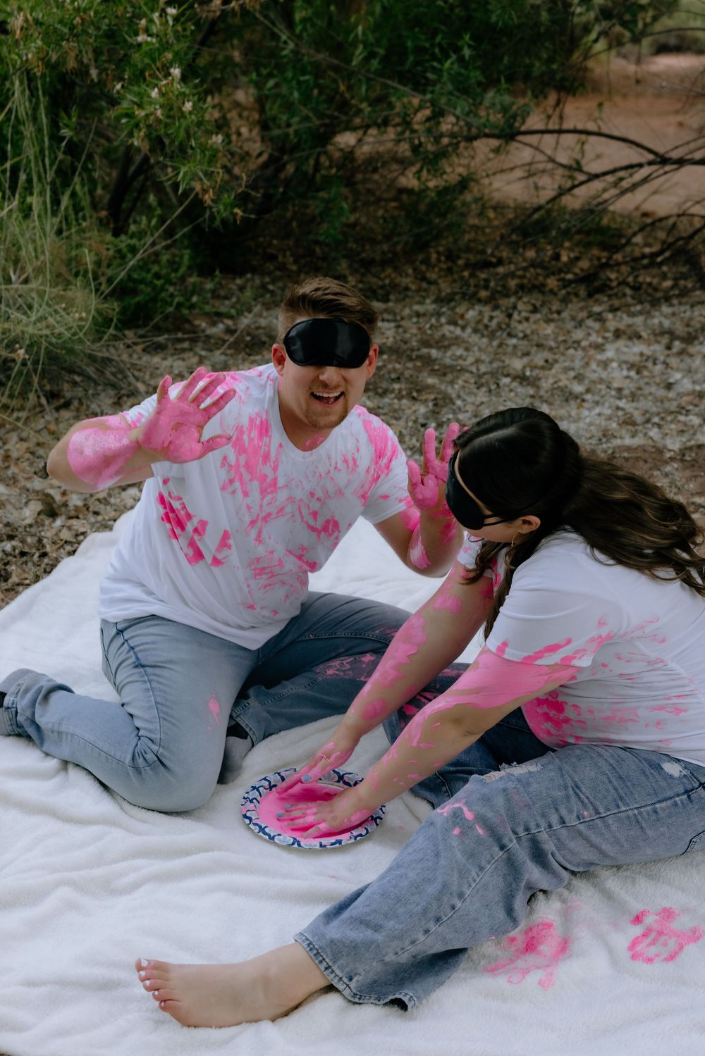 Two people wearing white clothes covered in pink paint sit on a blanket while playing with paint.