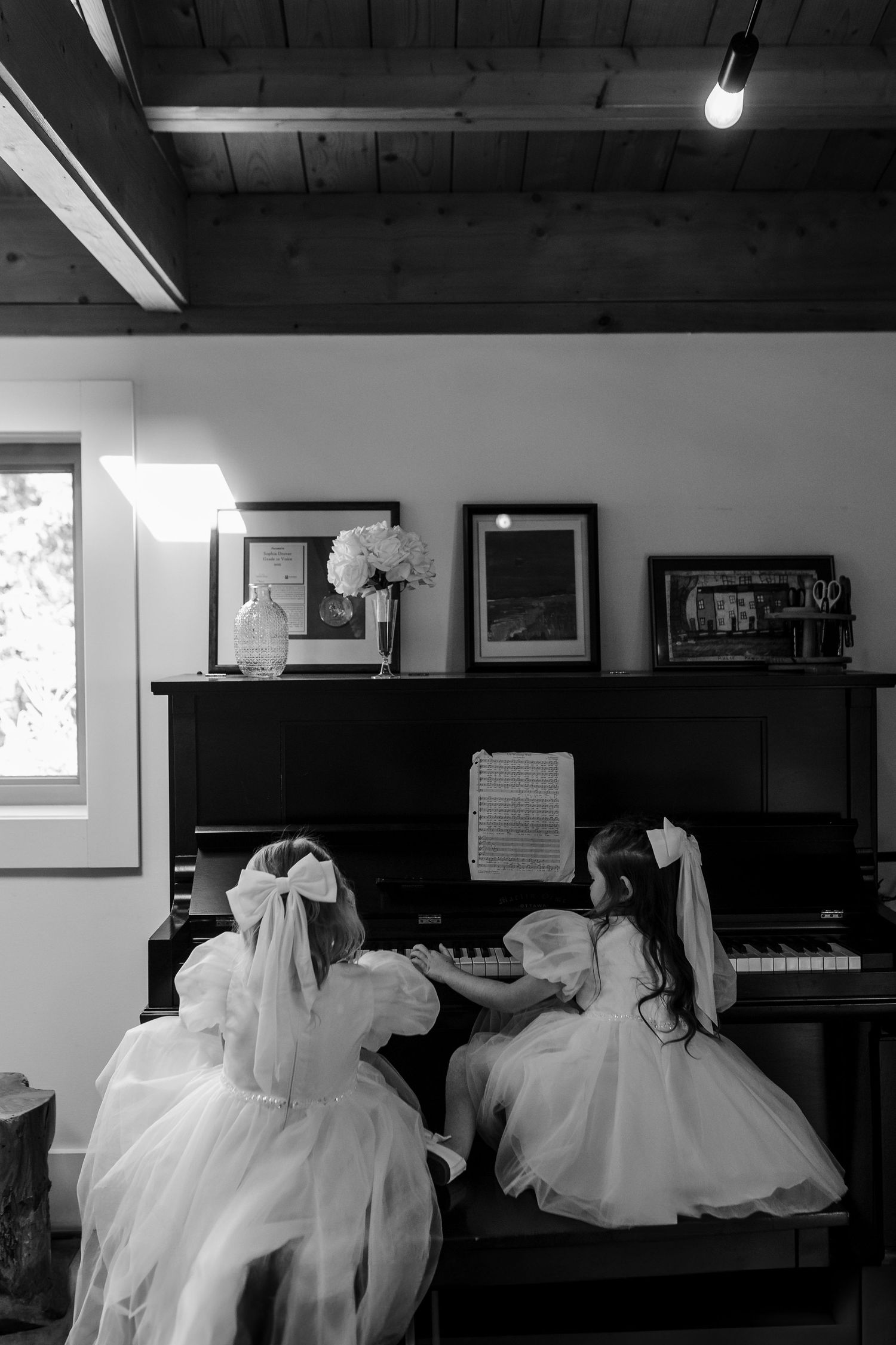 A black and white image shows white wedding dresses displayed in a room with a piano and framed artwork on the walls.