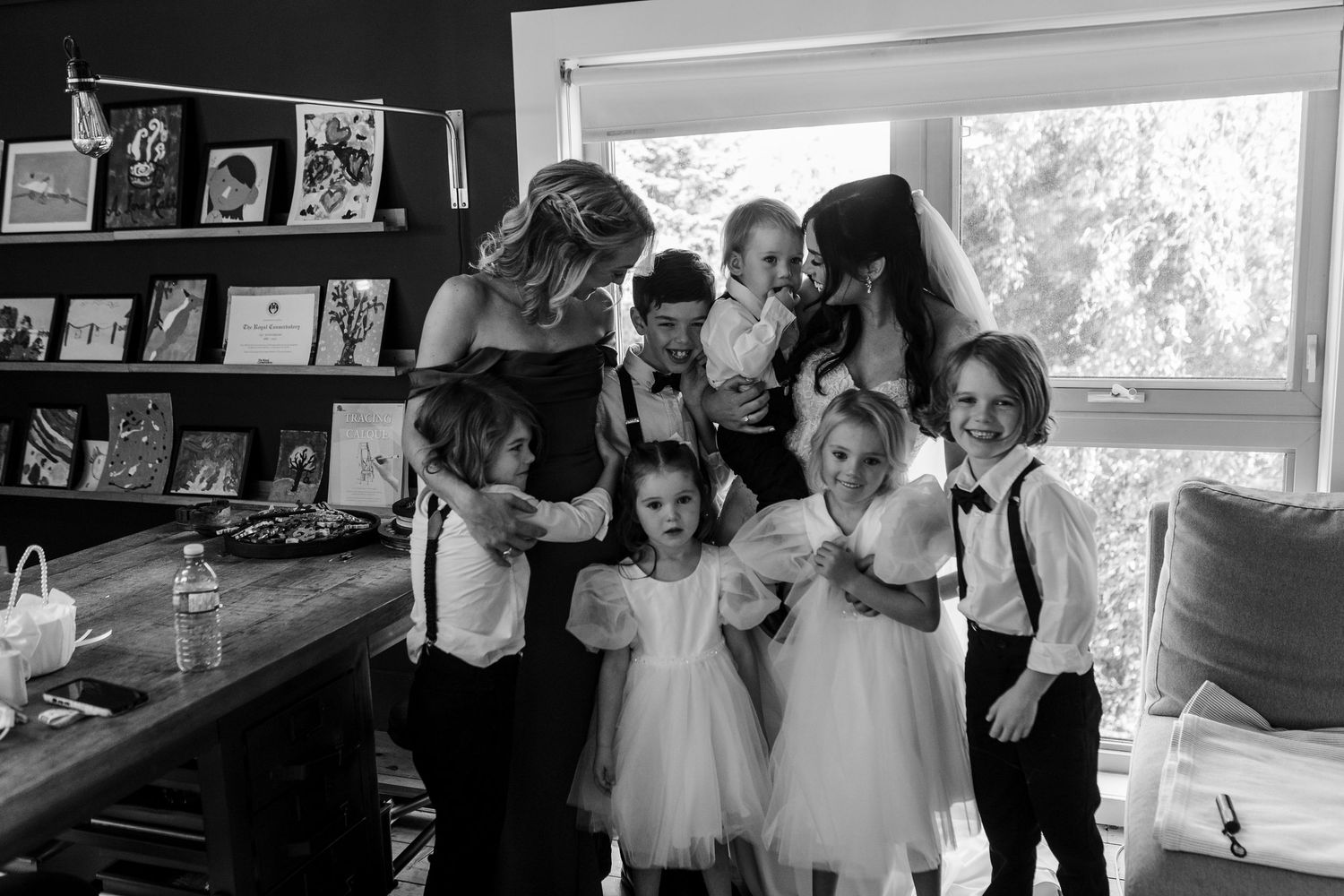 A black and white photo showing a group gathered together in a room with a window and photo display on the wall.