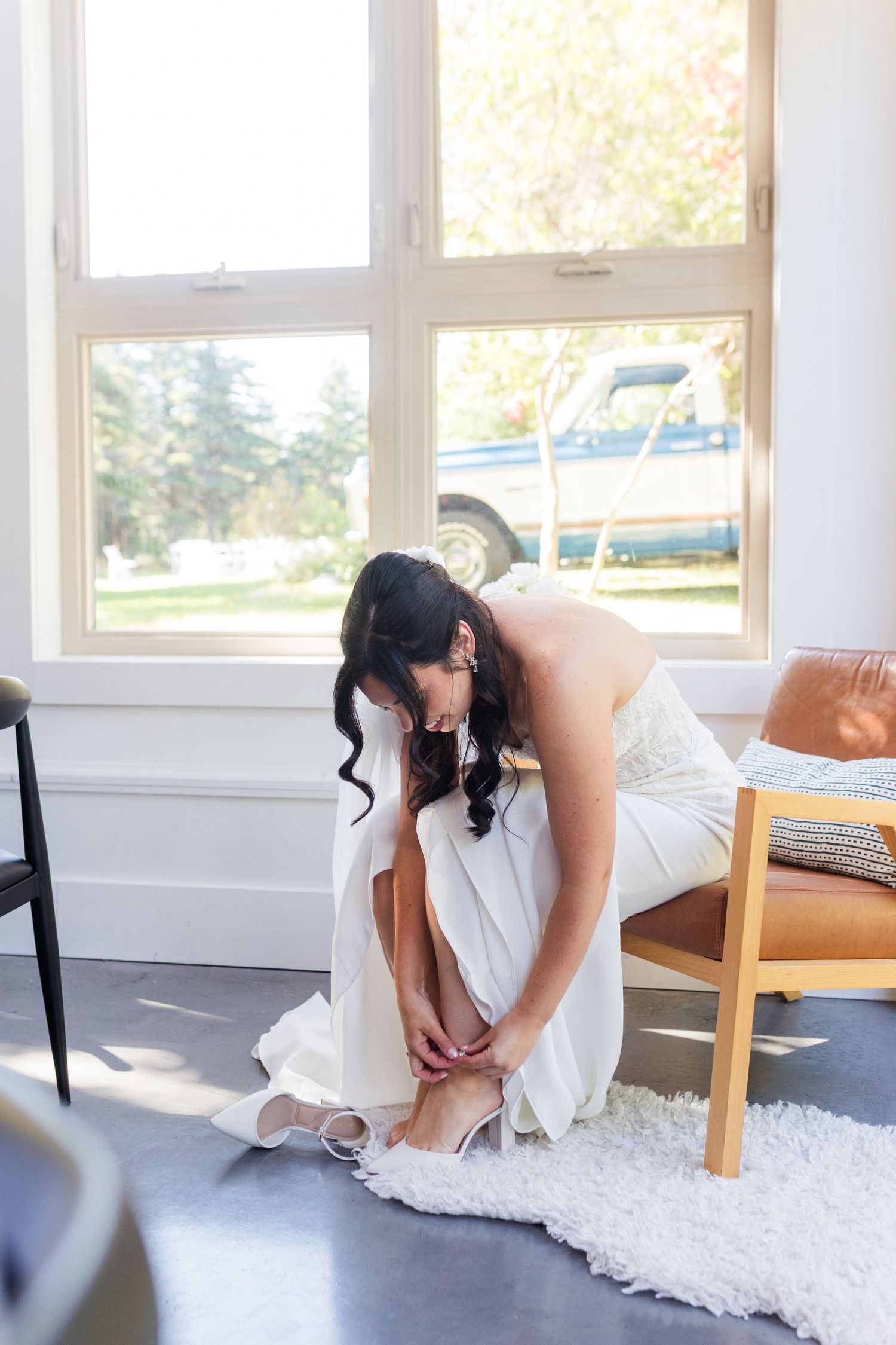 A person in a white dress sits on a fluffy white rug in a sunlit room with large windows.