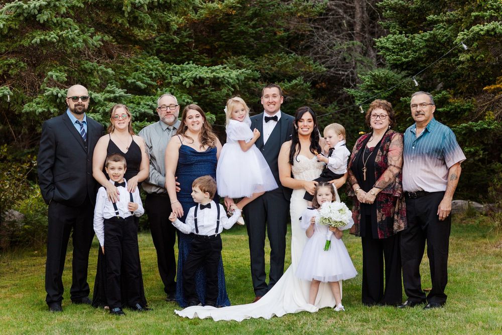 Wedding party members gathered outdoors for formal group photos on a grassy area with trees in background.