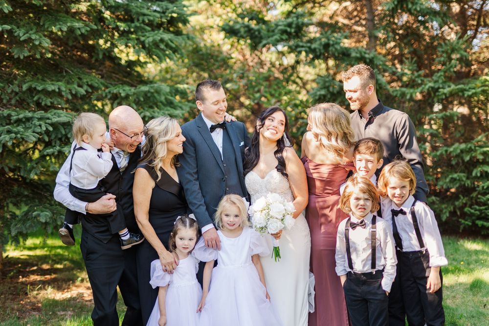 Wedding party members laugh and pose together outdoors in a garden setting with green foliage in the background.