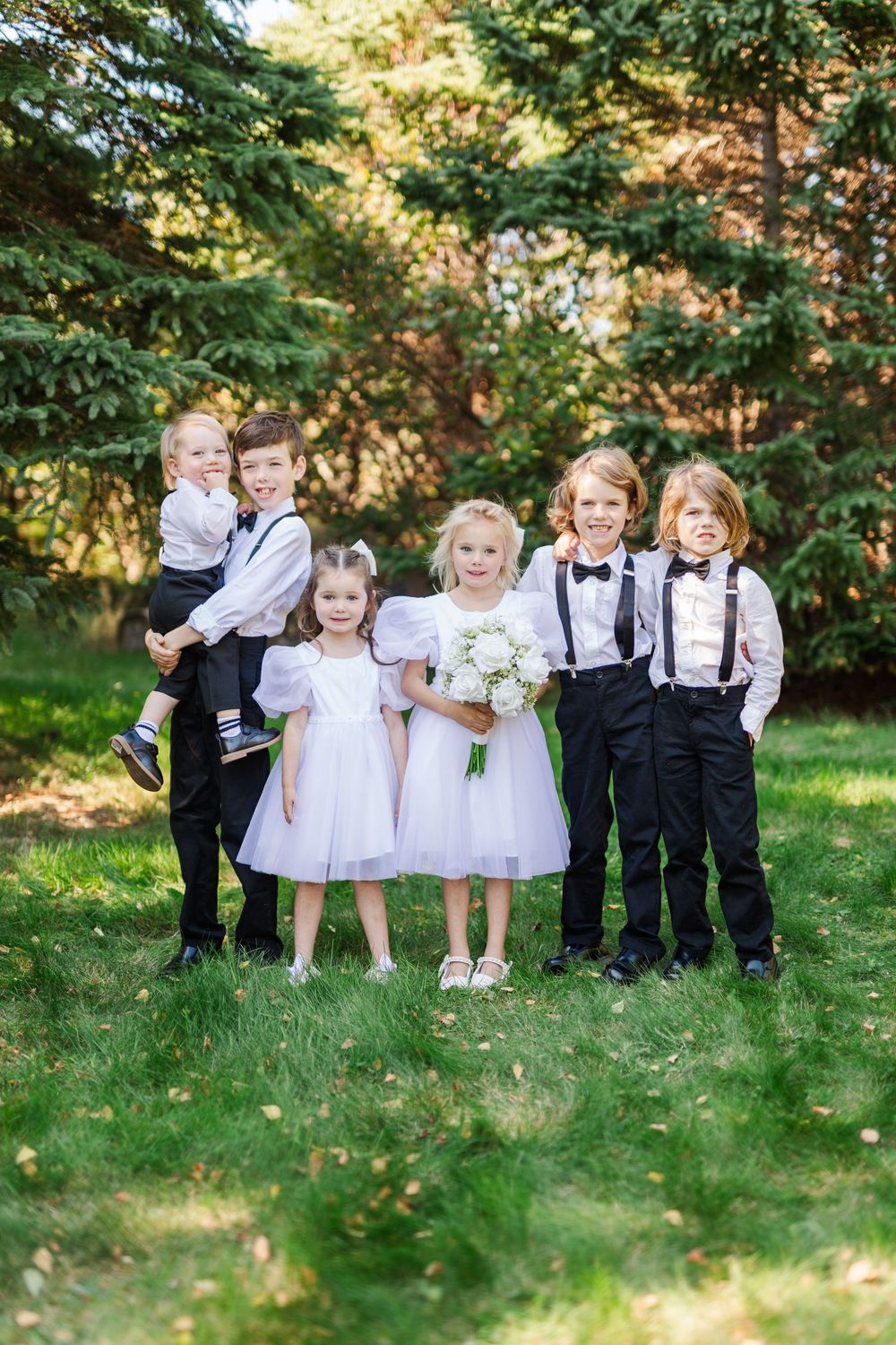 A group of children in formal attire playing and posing together on grass near evergreen trees.