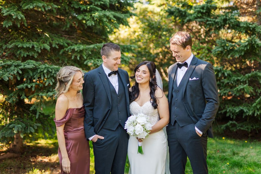 A wedding party of four people poses together outdoors with lush greenery in the background.