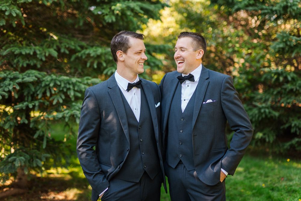 Two people in black tuxedos share a candid laughing moment outdoors during a wedding photoshoot.