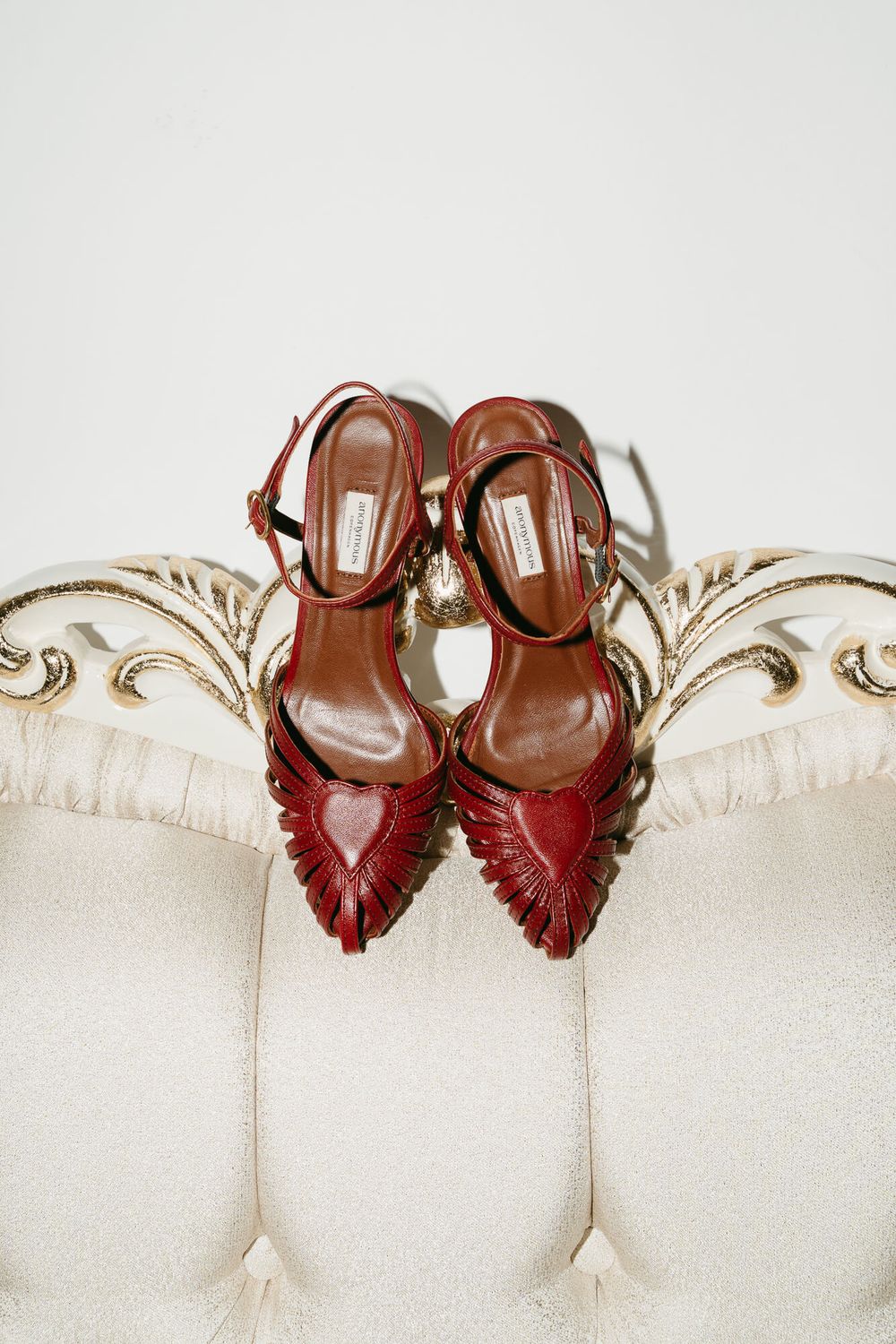 Elegant burgundy leather heels with ankle straps and pointed toes displayed on a cream-colored ornate settee.