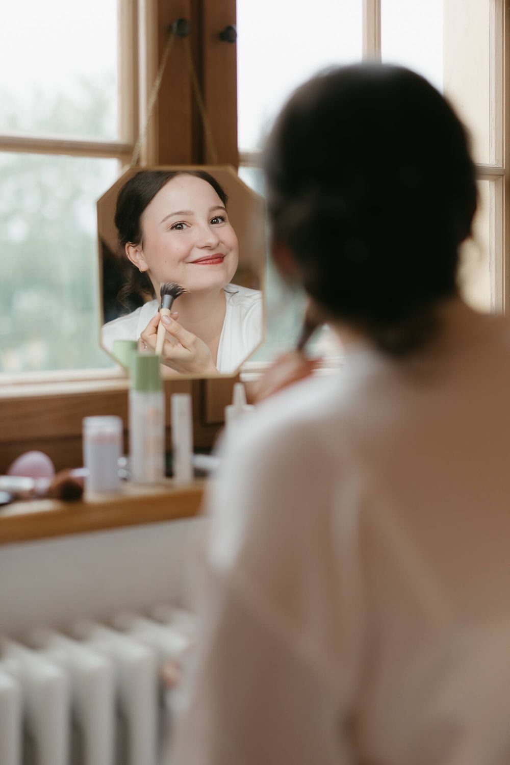 Someone applies makeup while looking in a mirror by a window with soft natural lighting and white decor.