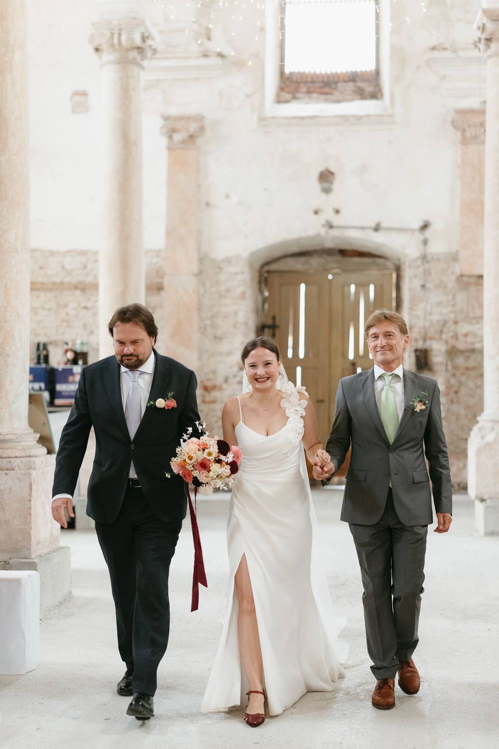 Wedding party walks joyfully together in a rustic indoor venue with exposed brick walls and white columns.