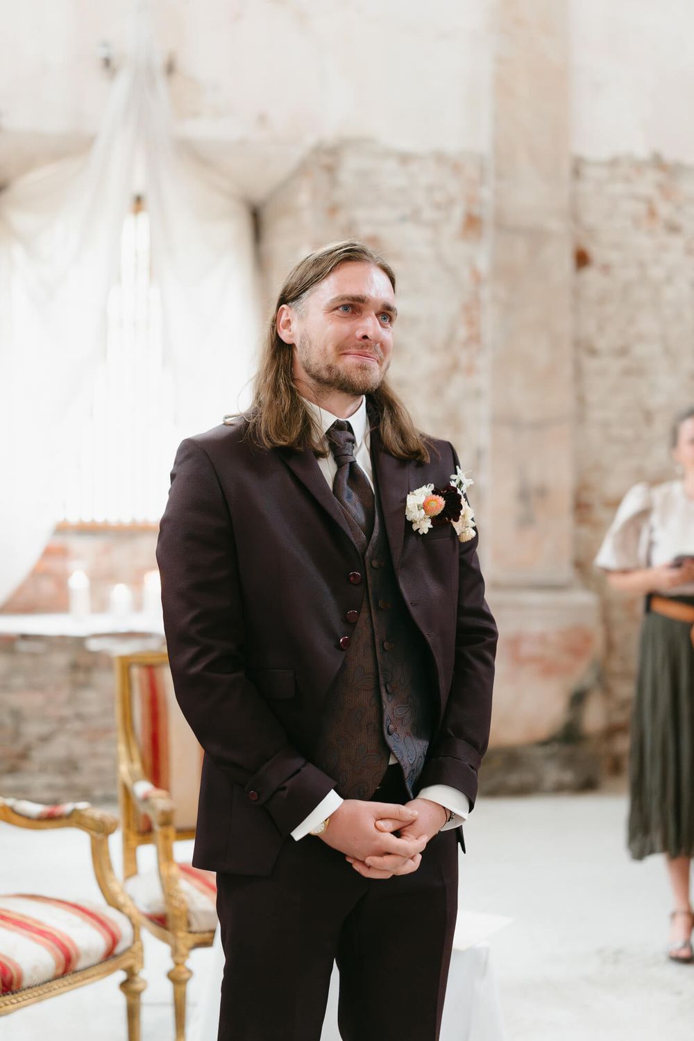 A groomsman in a black suit with a boutonniere stands at a wedding ceremony in a rustic brick venue.