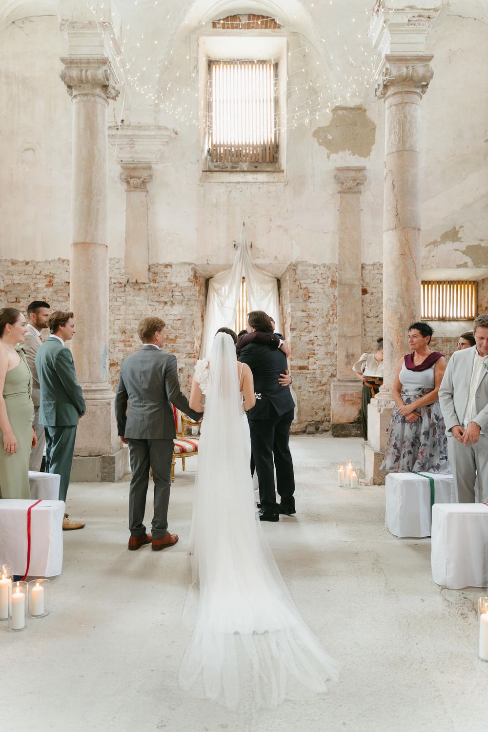 A wedding ceremony takes place in a rustic stone room with white chairs and romantic candlelight.