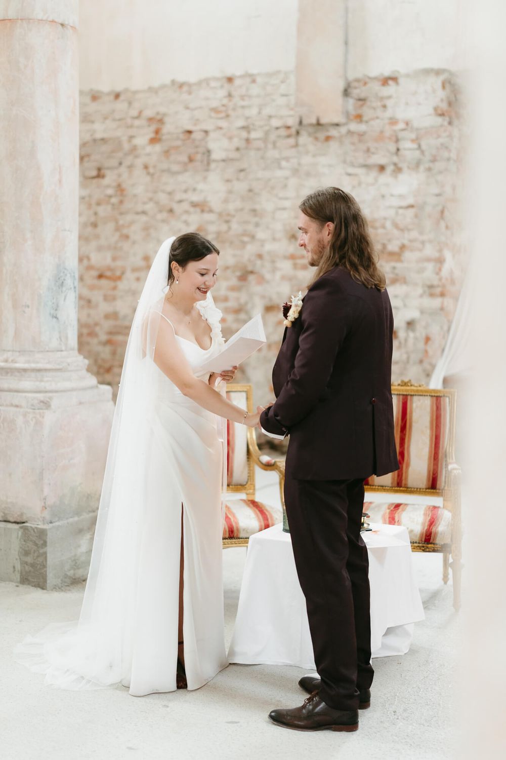 A bride and wedding party share joyful moments between white columns in a rustic brick venue.