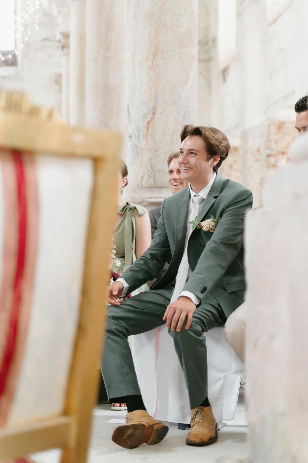 Groomsmen in green blazers sit casually on white columns in an elegant venue.