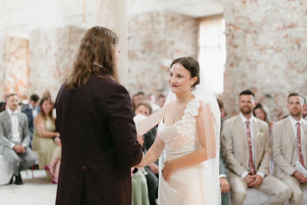 A wedding ceremony takes place in a rustic brick-walled venue with guests looking on as couple exchanges vows.