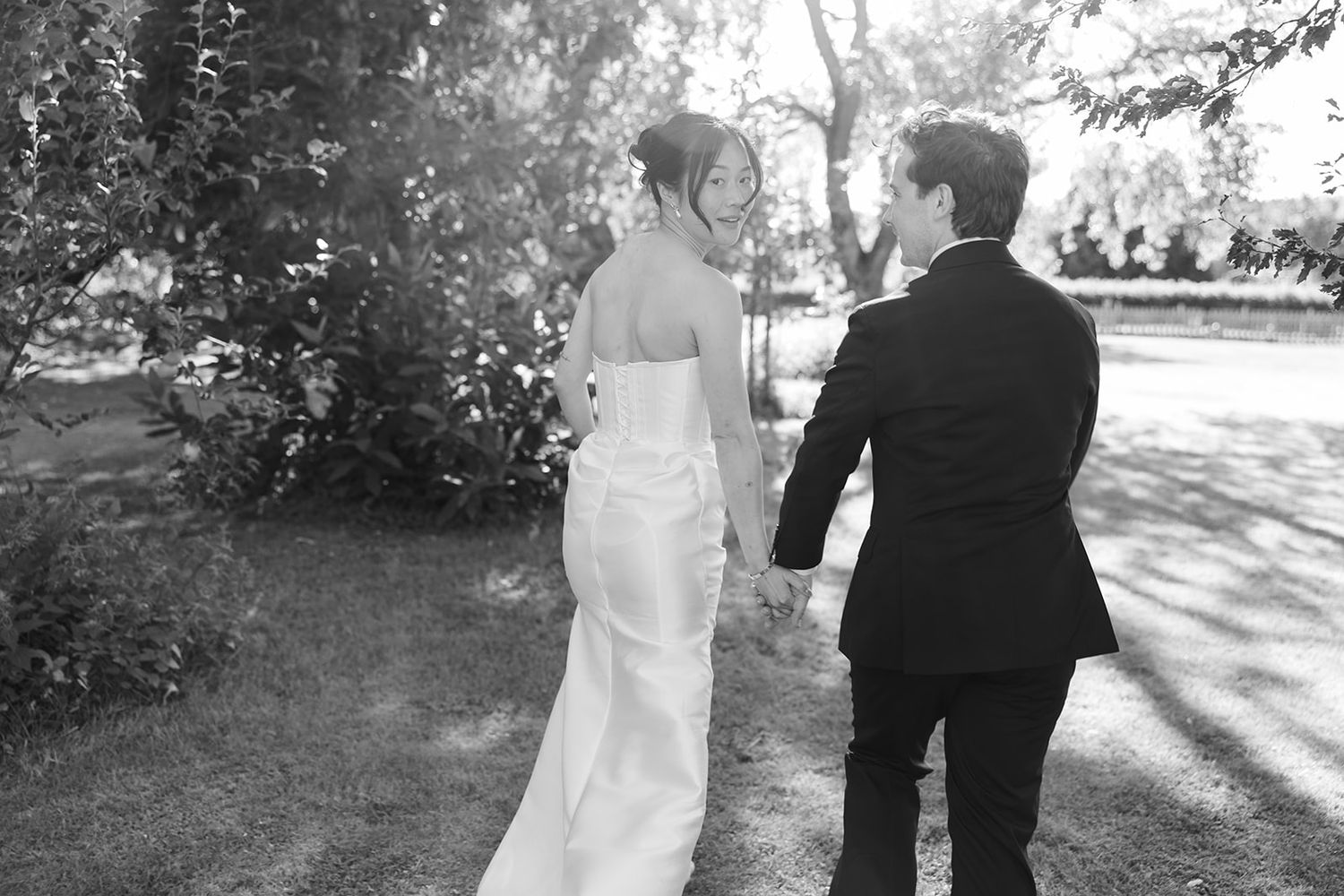 A couple walks through a sunlit garden path in formal wedding attire during an outdoor photo session.