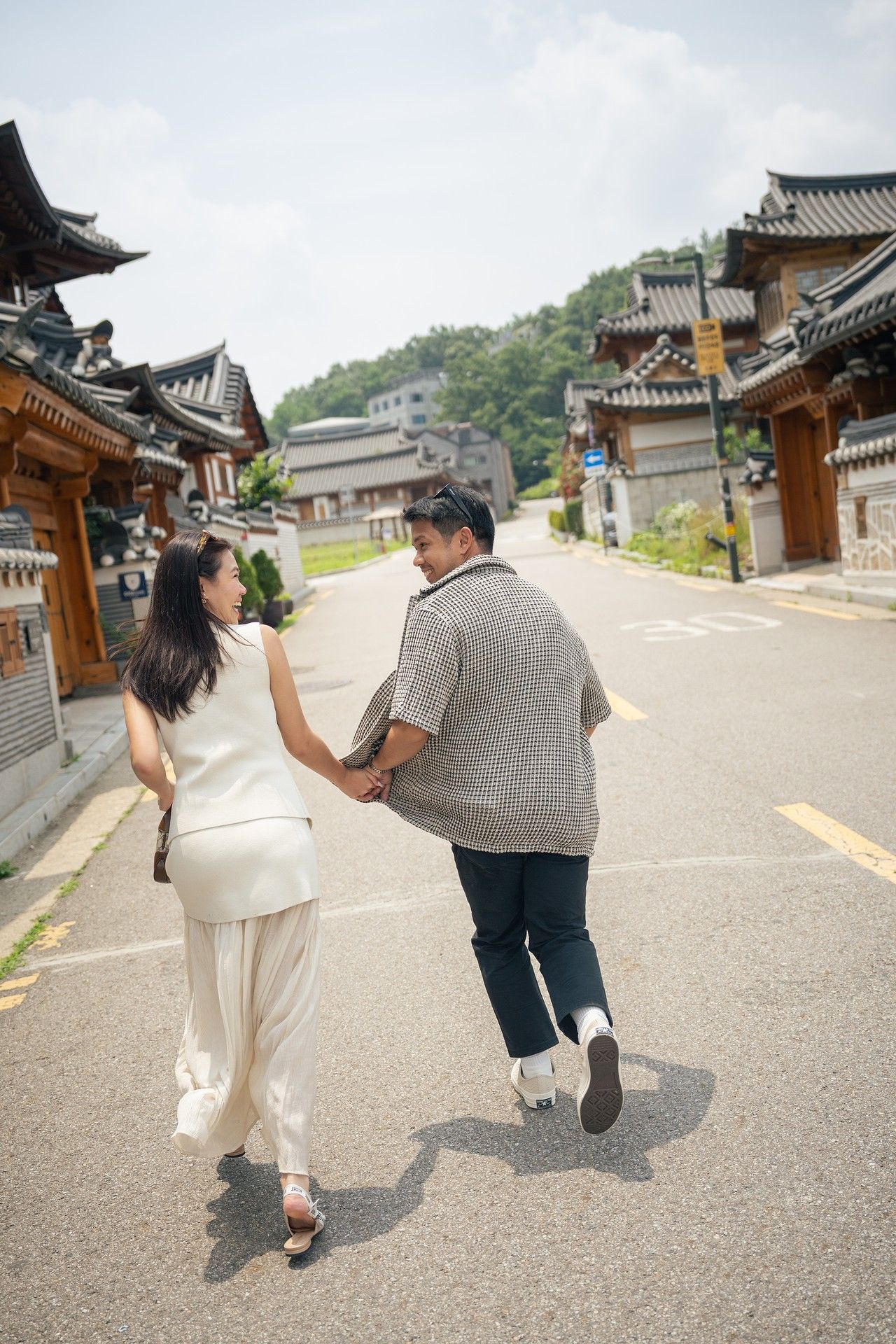 Two people playfully running and holding hands along a street lined with traditional Korean hanok buildings.