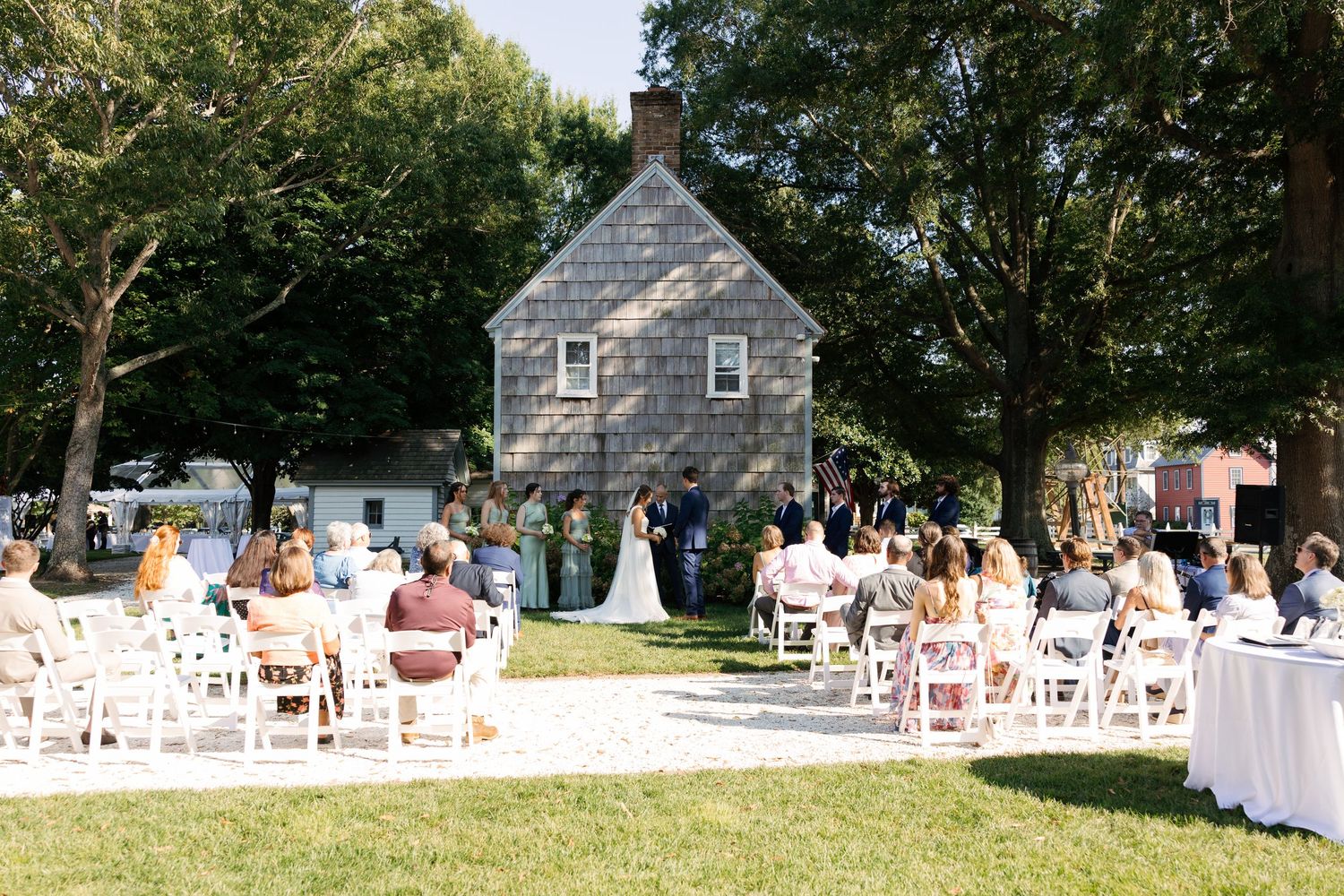 A panoramic view of an outdoor wedding ceremony setup with white chairs on a lawn near a stone cottage.