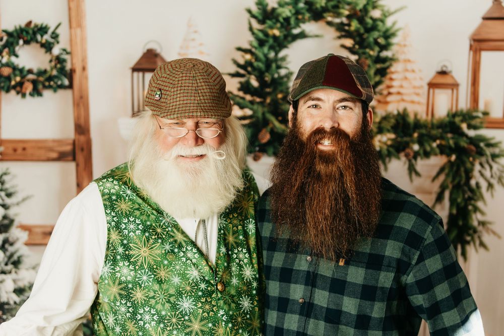 Two bearded individuals in holiday attire pose together with wreaths and lanterns in the background.